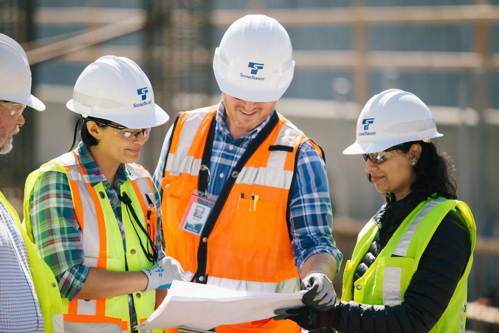 Four people in hard hats discuss a document out in the field.