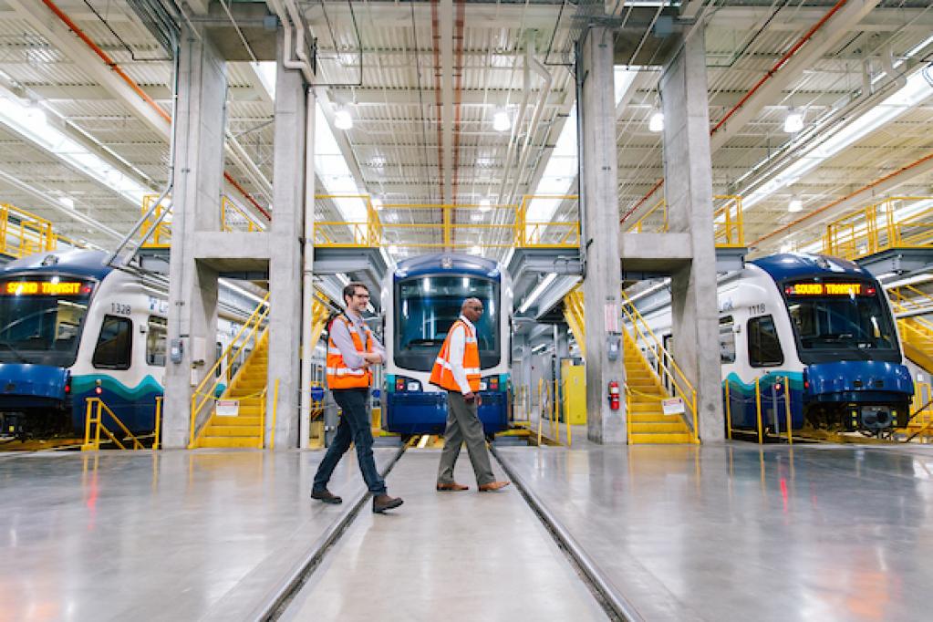 Two men in orange vests and hard hats walk past light rail vehicles at Sound Transit's Operations and Maintenance Facility.