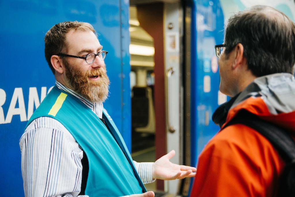 A man in a teal vest chats with another man by the door of a Sounder train.