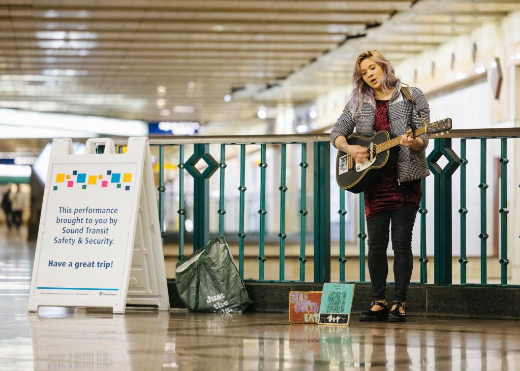 Ruby stands with her guitar at Westlake Station next to a sign that says 'brought to you by Sound Transit.'