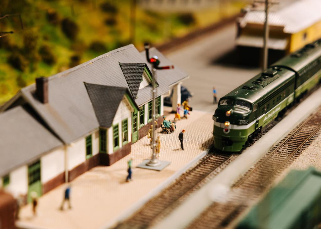 A green model train pulls into a station with tiny people waiting on the platform