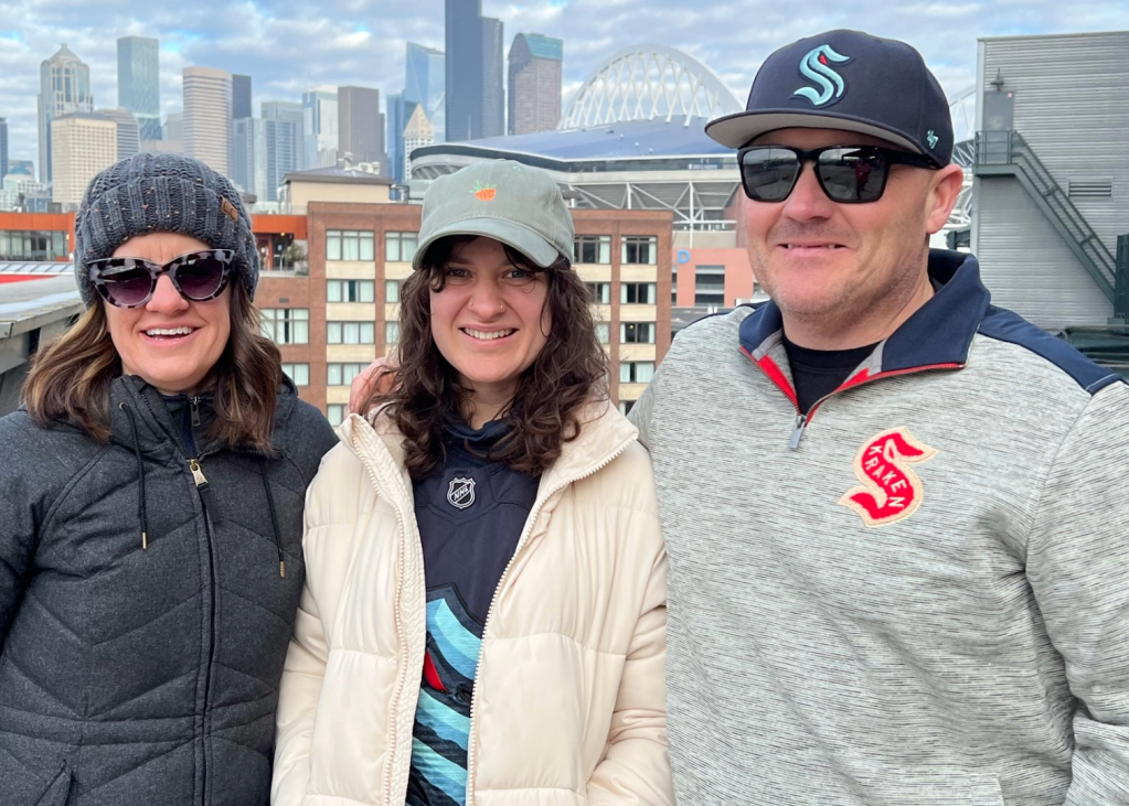 Three people smile with their Kraken gear on and downtown Seattle buildings in the background