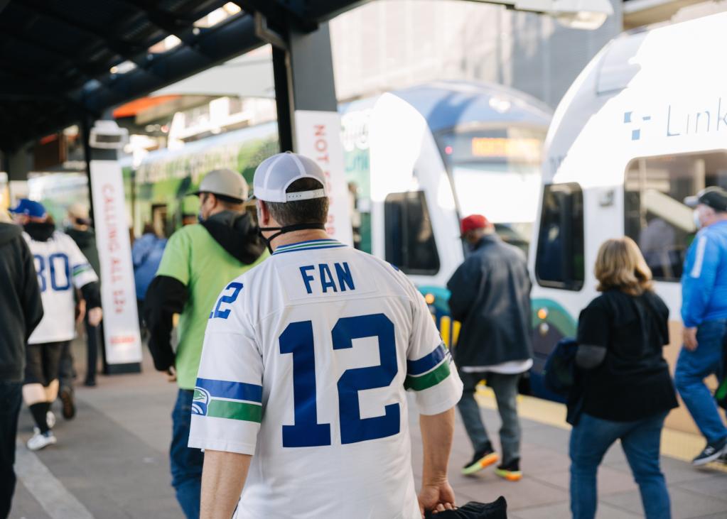 A Seahawks fan in a '12' jersey walks on a Link platform, with a train in the background