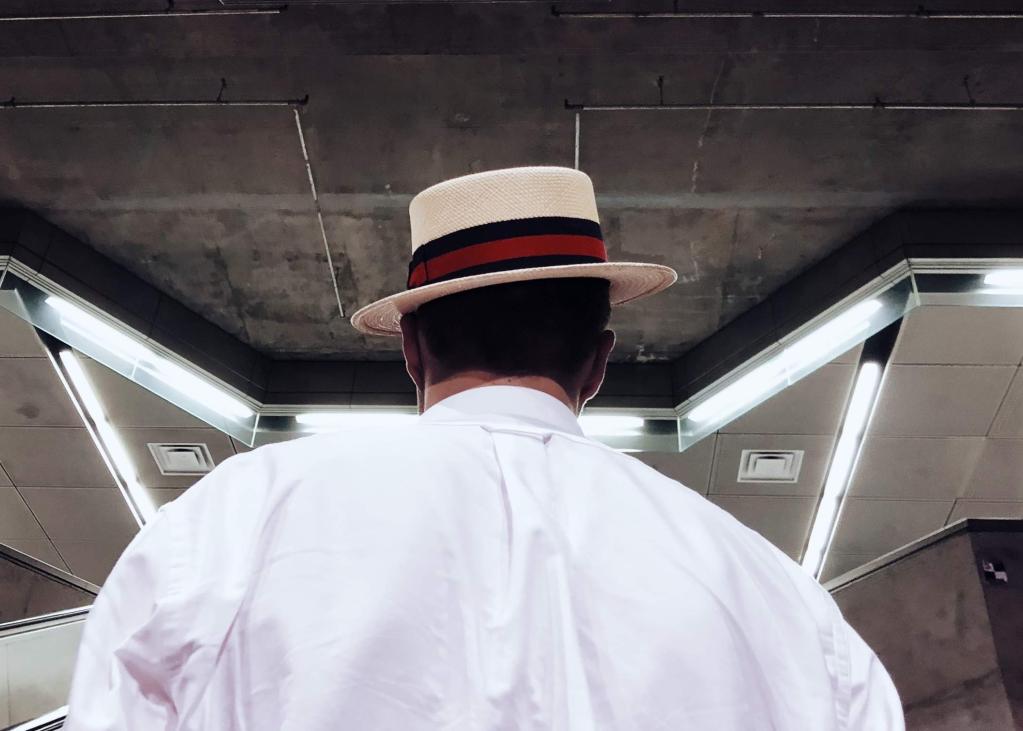 The back of a person's head as they sport a cool fedora while going up an escalator in a 1 Line station