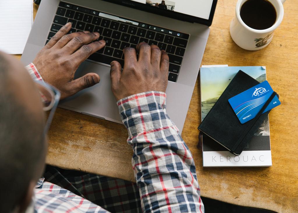 A person types on a laptop with a cup of coffee, notebook and ORCA card nearby on their desk.