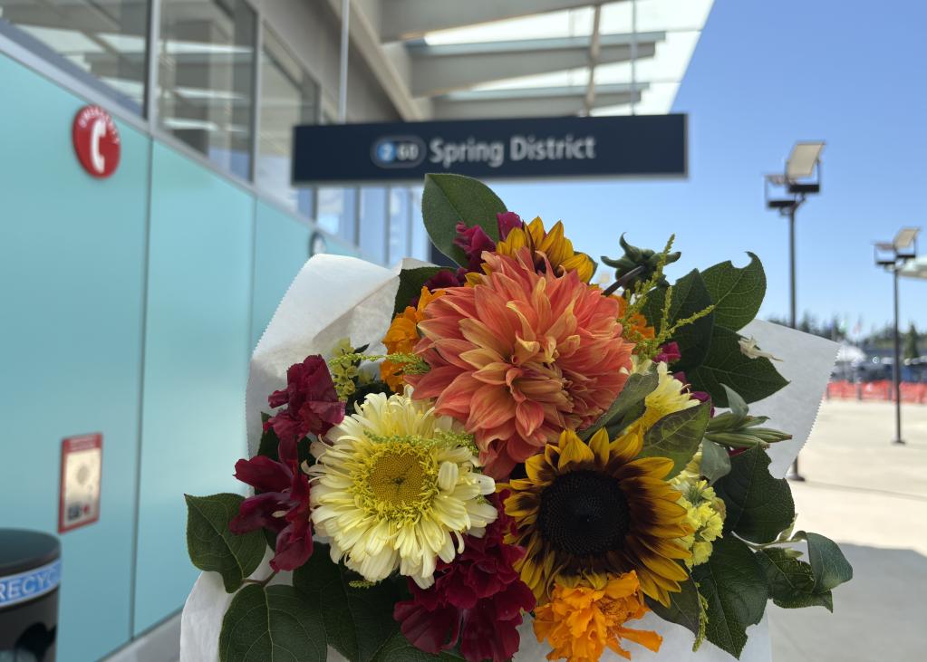 A bouquet of flowers, including pink dahlias and sunflowers, with a sign for the entrance to Spring District Station in the background.