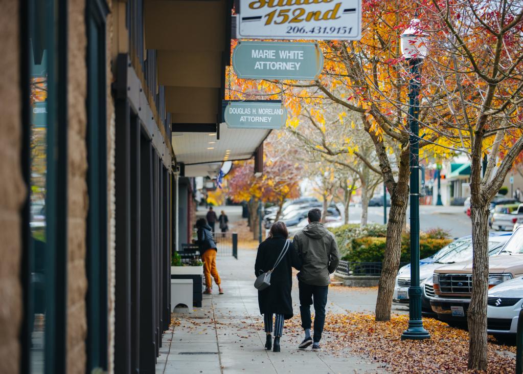 Two people walk on a sidewalk in downtown Burien amidst trees with orange and yellow leaves