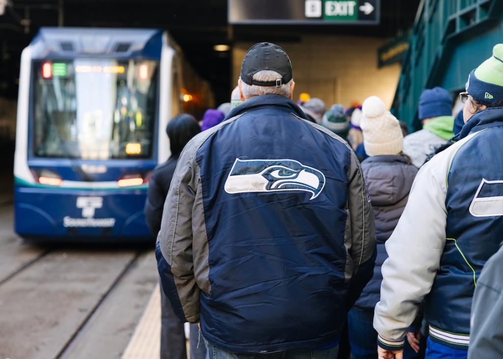 A Seahawks logo on the back of a fan's jacket. They are walking on the platform at International District/Chinatown Station, with a Link train in the background.
