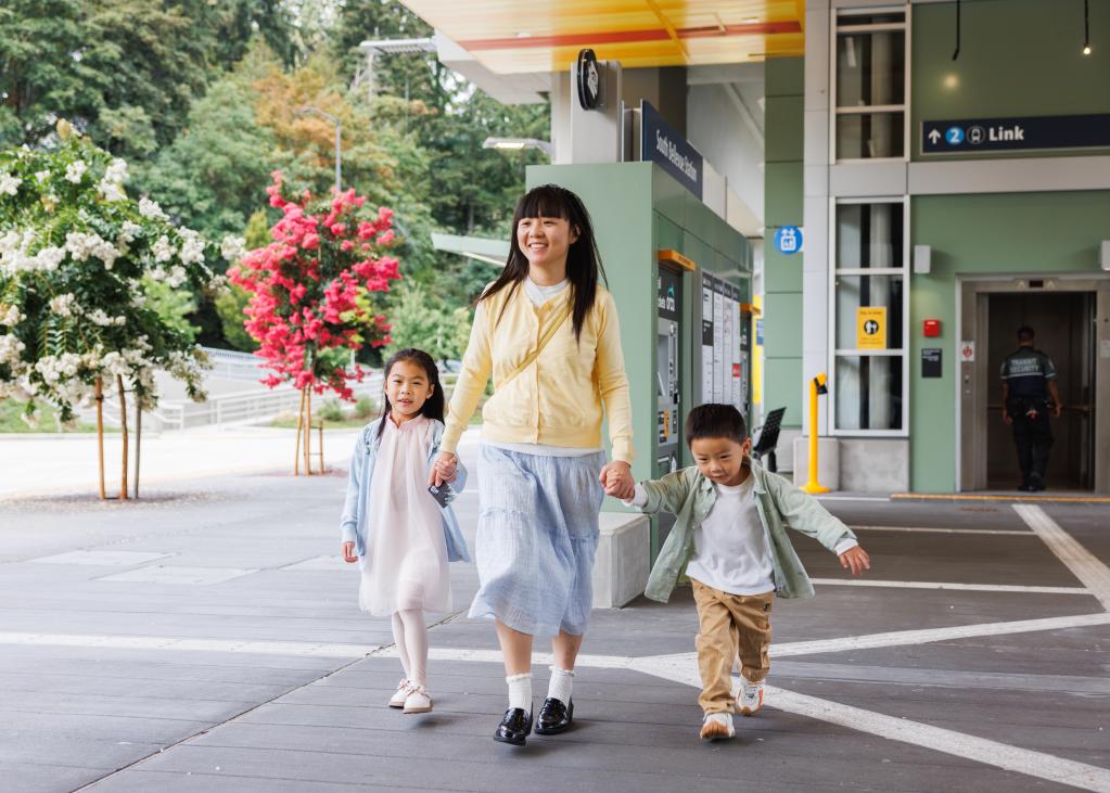 A mom and her two kids hold hands and walk near the entrance to South Bellevue Station on a sunny day
