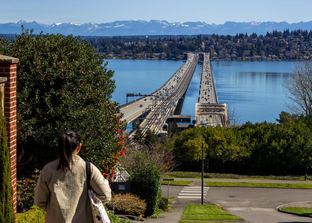 A woman with a ponytail walks on a sidewalk with a view of Lake Washington and the I-90 floating bridges