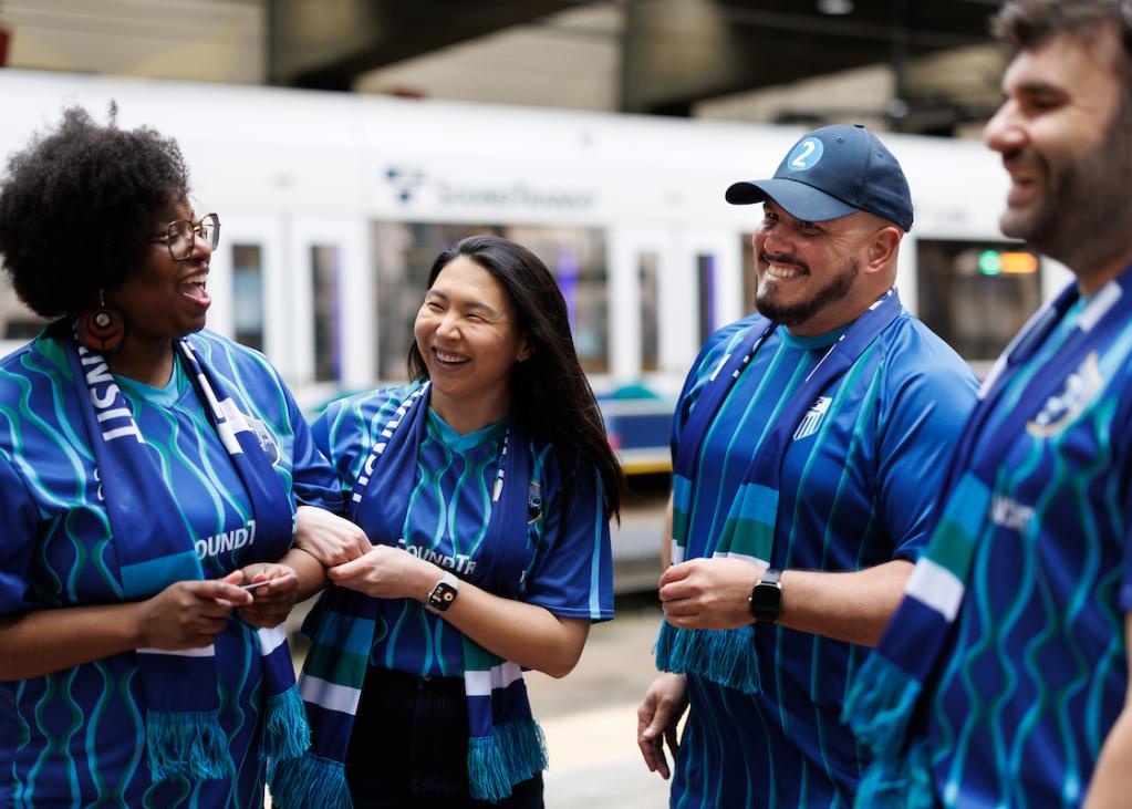 Four people in blue jerseys stand on the train platform at International District/Chinatown Station