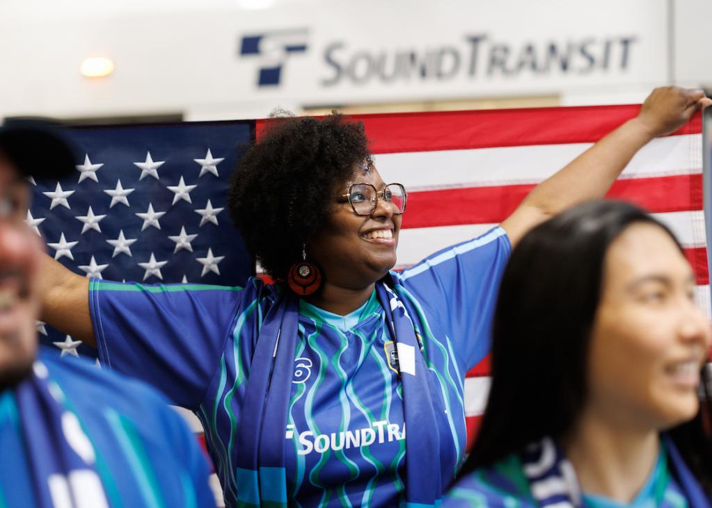 A soccer fan wearing a blue jersey holds a U.S. flag up behind her head. Two other fans are in the foreground of the photo