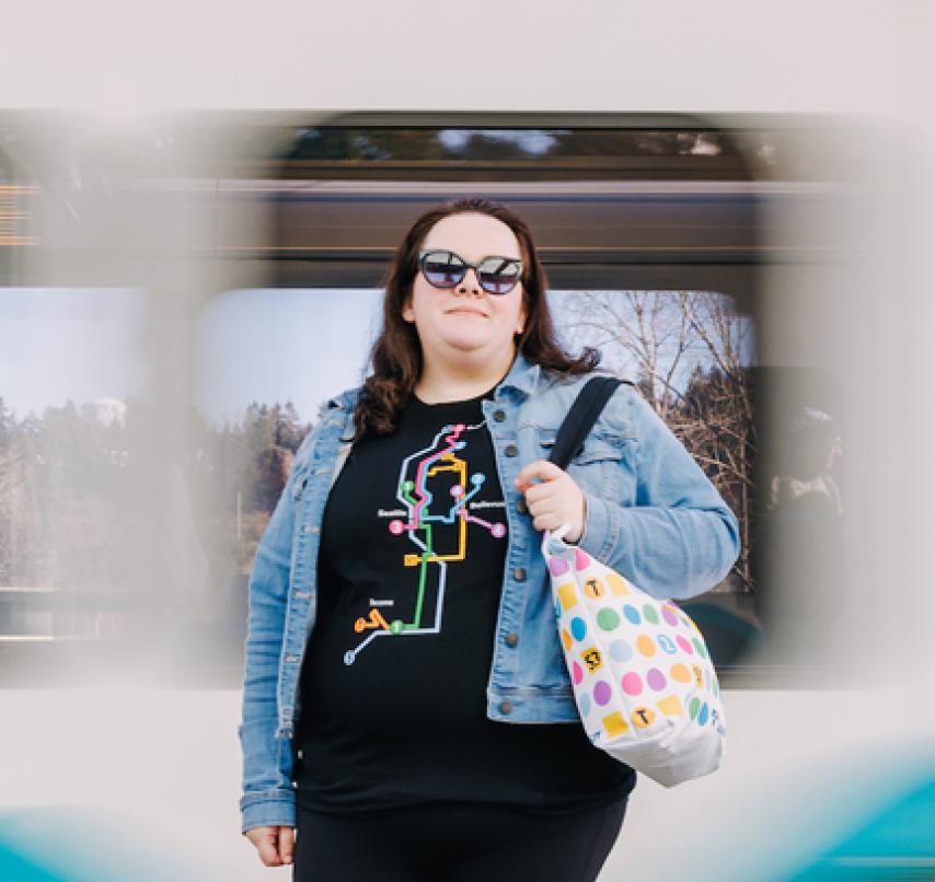 A passenger in Sound Transit shop gear stands on a station platform as a light rail train moves by.
